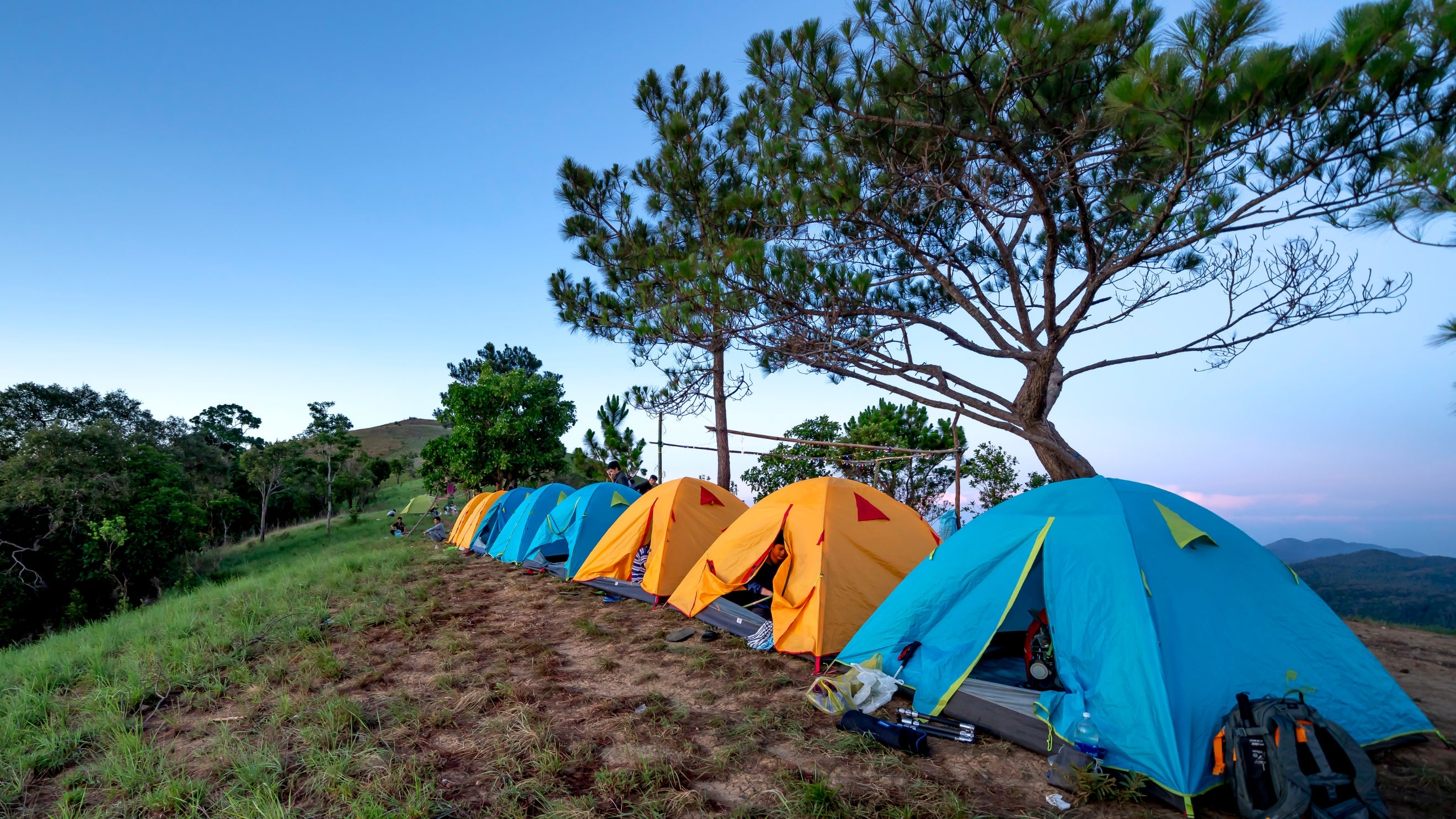 Row of colorful tents set up on a grassy hillside with trees and mountains in the background.