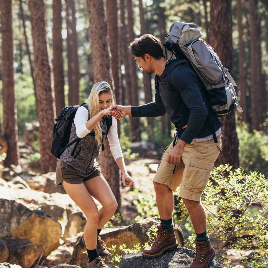 Two hikers in a forest, one helping the other climb over rocks.